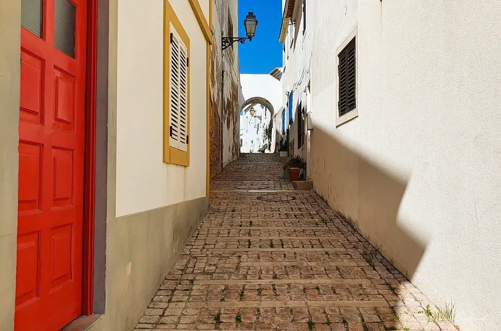 Narrow cobblestoned street between cute houses leading to a white arch