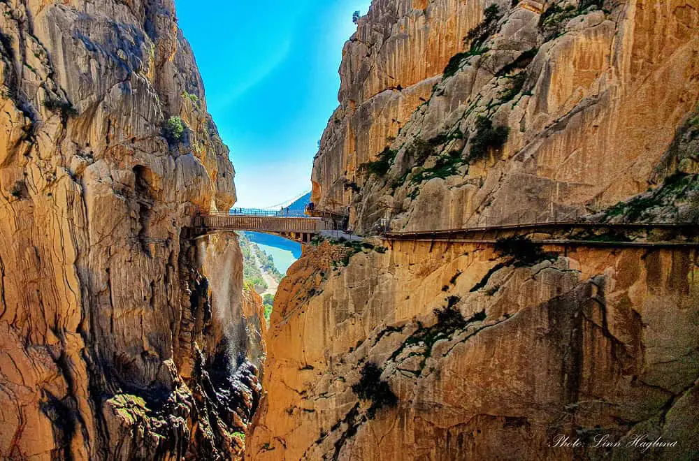 A wooden path along the vertical cliff wall leading to a hanging bridge across a narrow gorge at El Caminito del Rey hike in Spain.