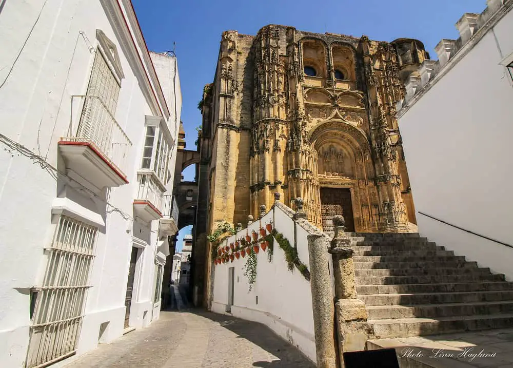 The beautiful church with intricate details in stone of Minor Basilica of Santa María de la Asunción in Arcos de la Frontera, standing on top of a set of stairs and connected with an arch with the whitewashed houses on the other side of a narrow street.