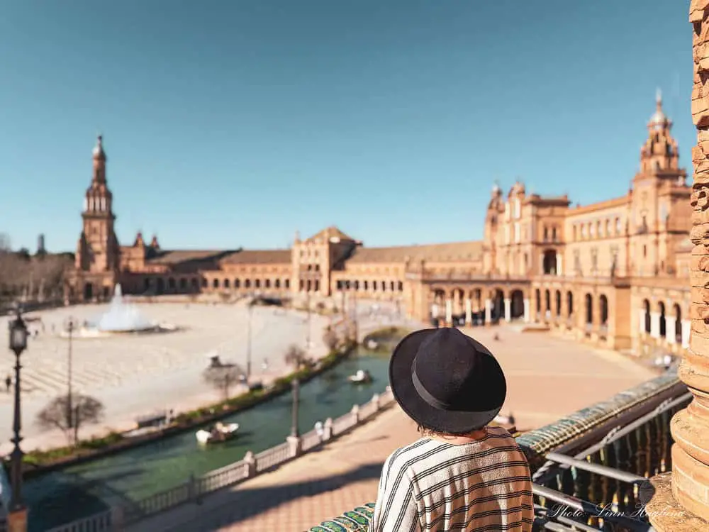 Me, with a black, wide-brimmed hat on a balcony in Plaza de España in Seville, Spain, looking at the square on a sunny day. Visiting Plaza de España is one of my favorite free things to do in Seville.