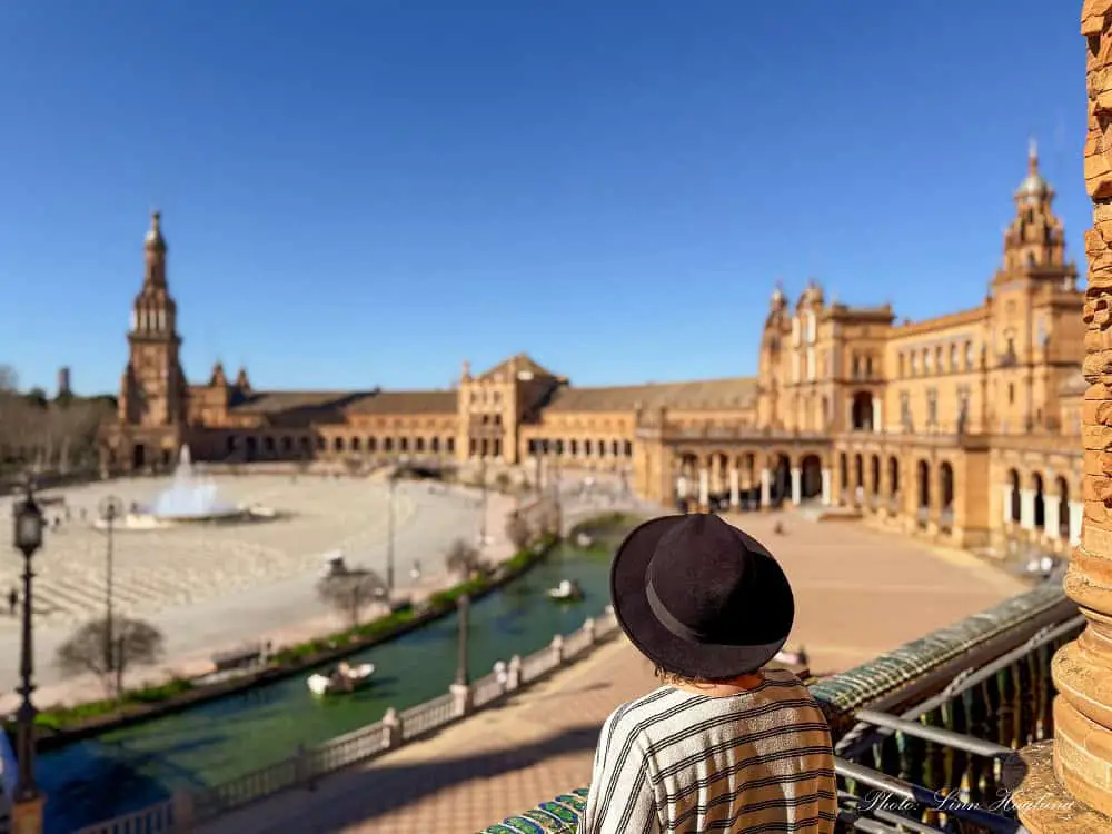 Me, with a black, wide-brimmed hat on a balcony in Plaza de España in Seville, Spain, looking at the square on a sunny day. Visiting Plaza de España is one of my favorite free things to do in Seville.