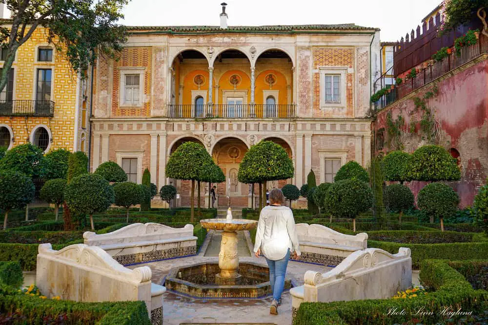 Me, a light-colored shirt and jeans, walking by a beautiful stone fountain in a perfectly groomed, green garden in Casa de Pilatos in Seville Spain. In the background is a yellow and white house with tall arches over two floors.