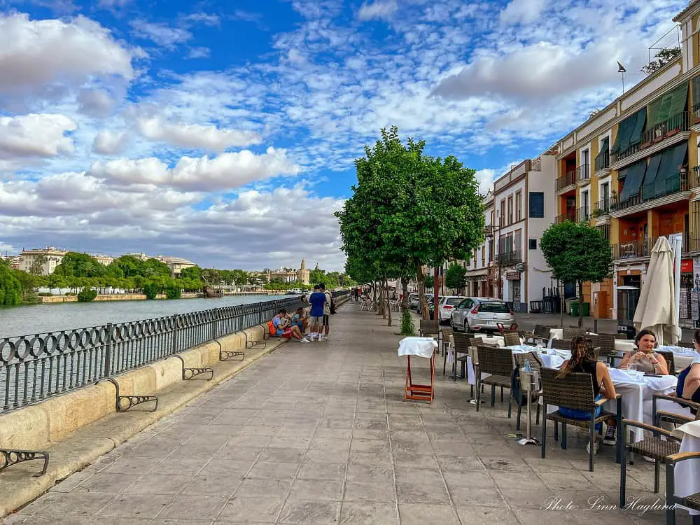 People eating and drinking in busy bars in Calle Betis along the Guadalquivir River in Seville Spain. The sky is blue, dotted bith white clouds, and the views of the other side of the river include the Torre del Oro.