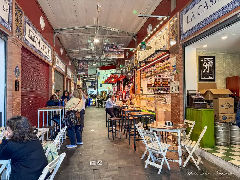 People eating tapas and drinking at tables in Triana Market in Seville Spain. Some stalls are closed, but the ones that are open are busy and decorated with Andalusian tiles.