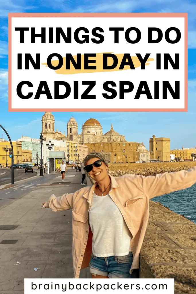 Me, with a pink jacket and sunniesopening my arms and smiling in front of the views of Cadiz Cathedral on the promenade of Cadiz Spain.