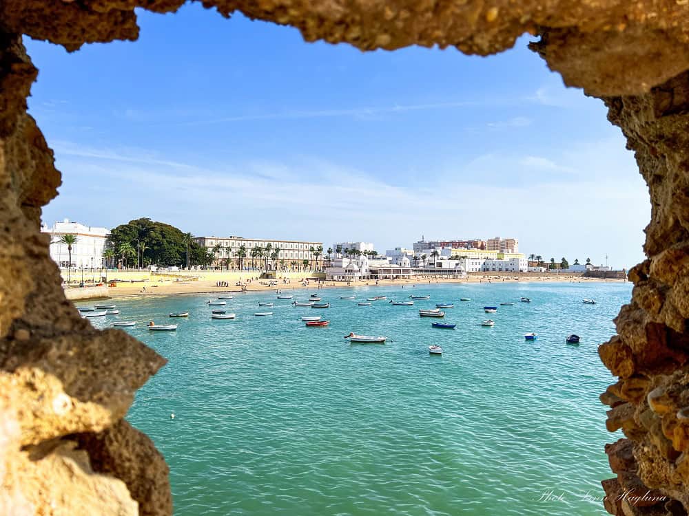 Scenic view of La Caleta Beach in Cádiz, Spain, framed by a stone window inside Santa Catalina Castle, overlooking golden sand, blue ocean, and historic coastal landmarks.
