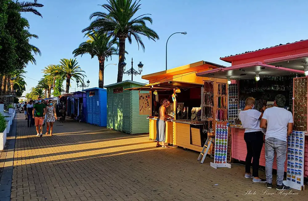 A pedestrian street lined with small colorful, wooden stands where people shop for handicrafts and other items. There are palm trees and the sun is low on the sky making the shadows long.