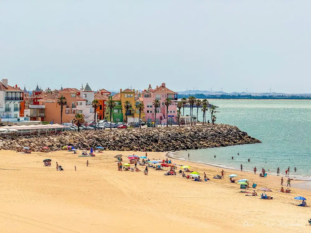 The colorful Puerto Sherry in El Puerto de Santa Maria overlooking a beautiful beach. People are swimming in the sea and colorful parasols dot the beach where people are sunbathing.