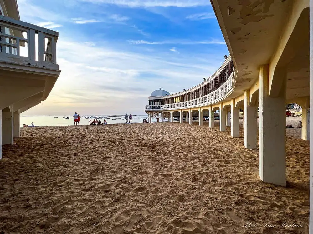 A long building elevated with poles, leading towards the water in La caleta Beach in Cadiz city at sunset. This is a favorite beach near Seville among locals who prefer an urban beach setting with things to do nearby.