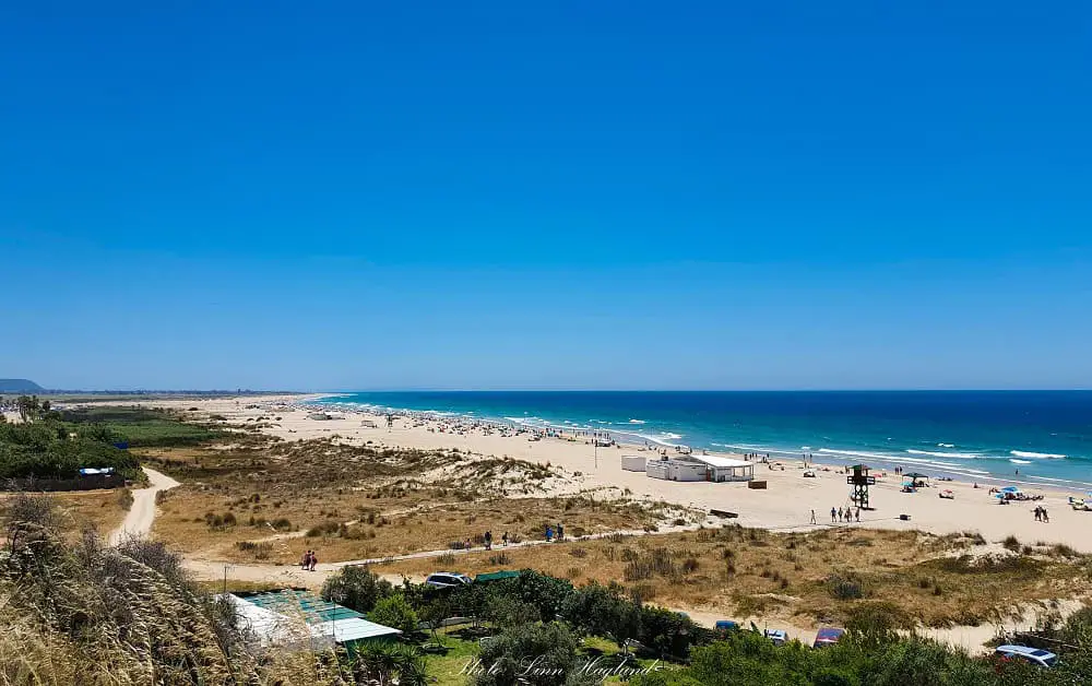 Conil de la Frontera Beach, with people walking and sunbathing on the beach, and others swimming in the blue sea on a sunny day. There are long boardwalks from the parking area and main street crossing a wide area of dunes to the beach.
