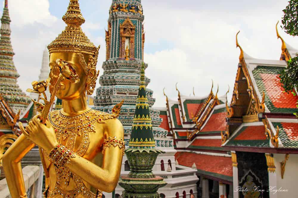 A golden Buddha statue with colorful rooftops of the Grand Palace in the background