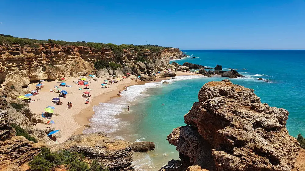 People sitting by their colorful beach umbrellas on Calas de Roche Beach, which is wrapped in cliffs. This is one of the best beaches close to Seville that are in a completely natural setting.