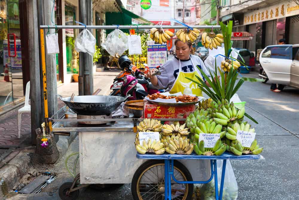 A woman selling street food and fruit from a stall on a street in Bangkok