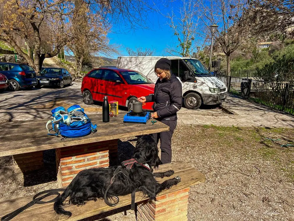 Spain van life, me and my dog Ayla cooking coffee at a picnic table next to parked cars and campervans.
