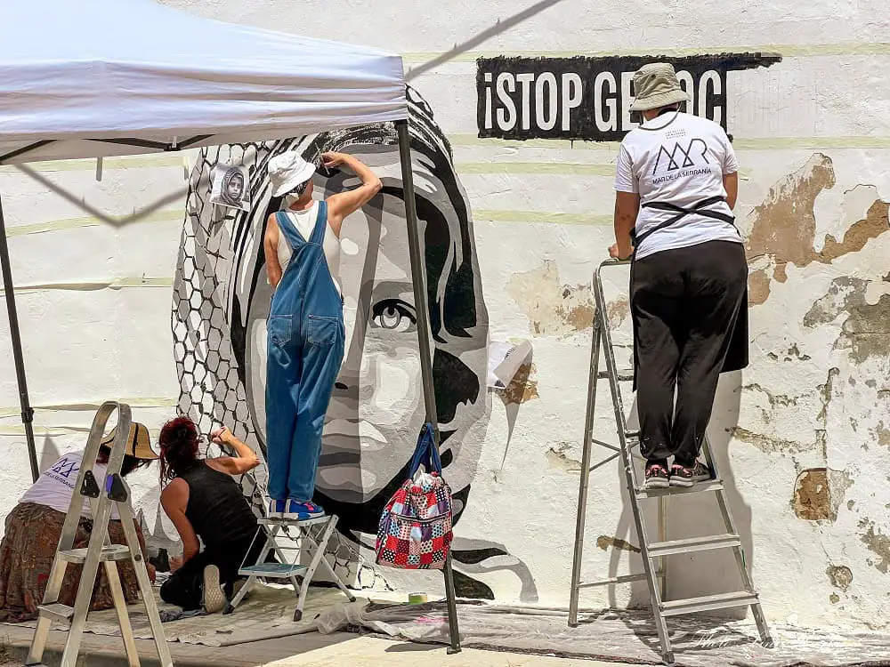 Street artists painting a girl on a wall with the text "Stop Genocide"