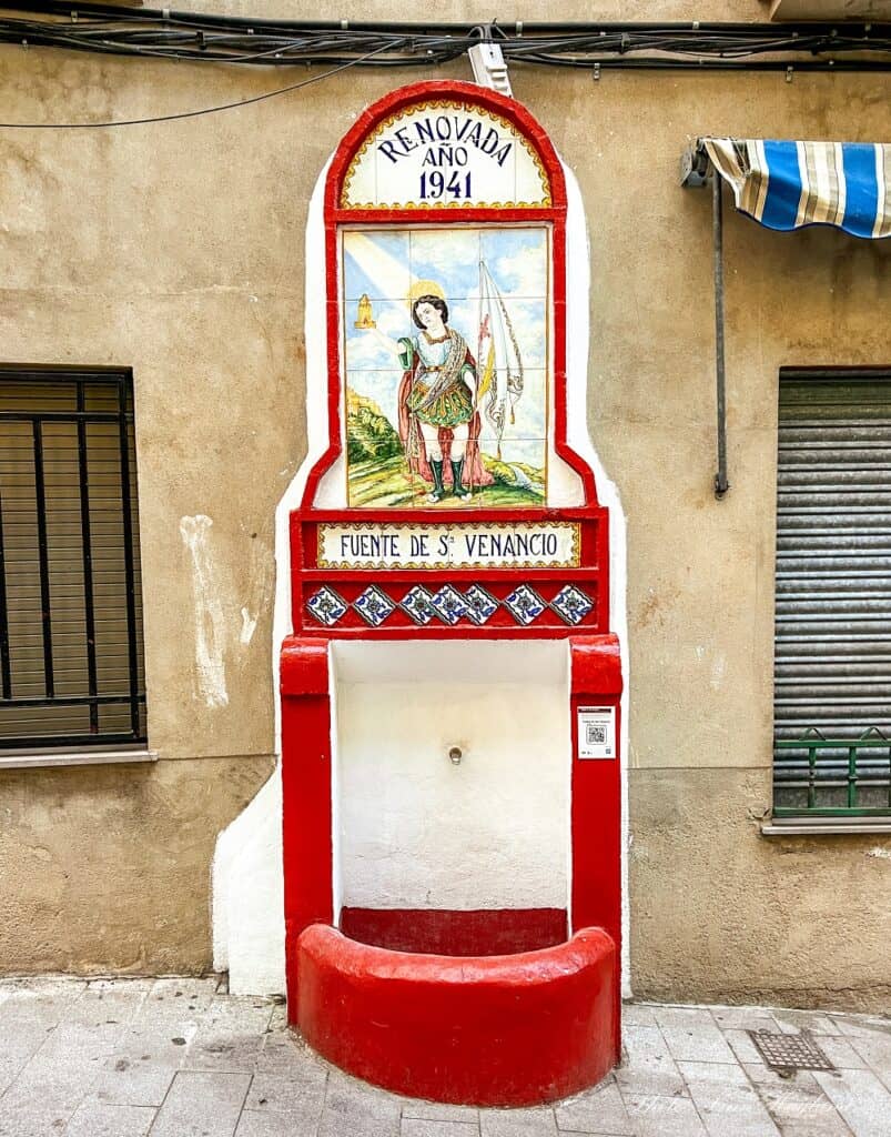 Water fountain dedicated to St. Venancio which is red painted and has a painting of the saint, saying it was renovated in 1941.