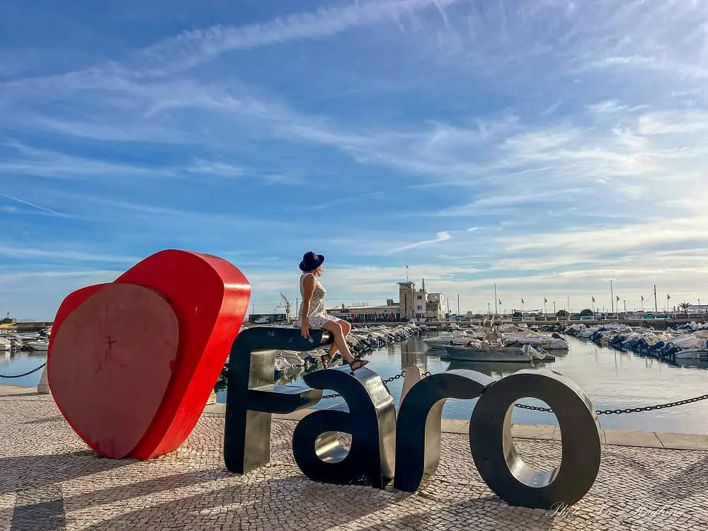 Me sitting on the I love Faro sign, looking at what to do in Faro Portugal marina, as boat trips go past.