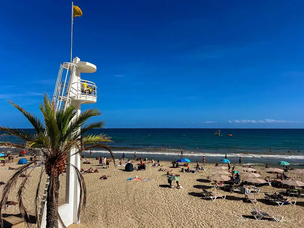 The beach in Alicante with people sunbathing and a lifeguard tower overlooking the beach for security which is one of the top reasons to visit Alicante Spain.