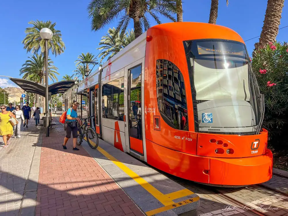 A red tram in Alicante Spain with a woman boarding with her bike.