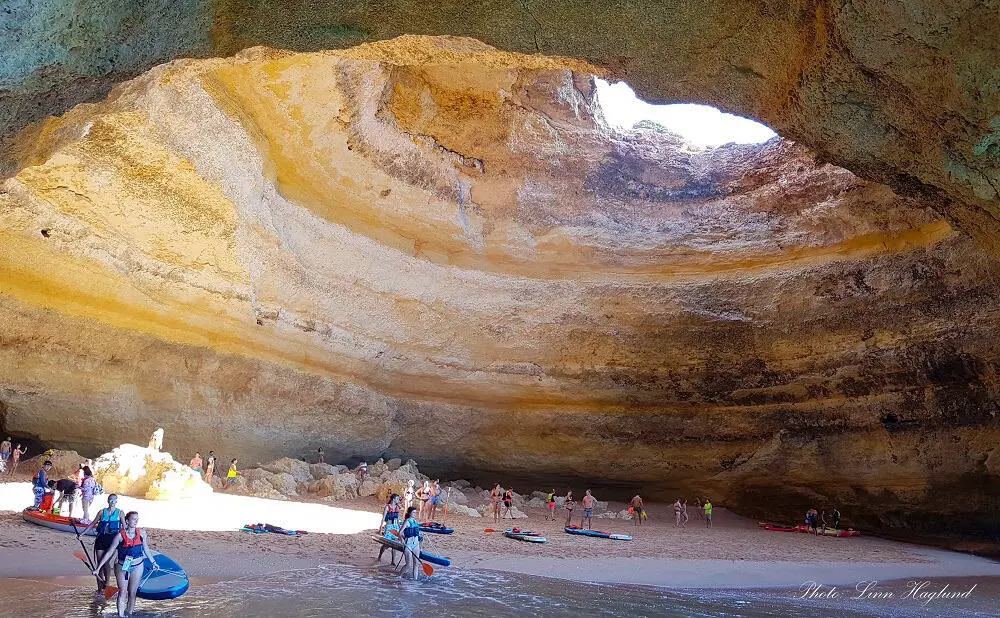 People with kayaks and paddleboards inside Benagil Cave in Algarve Portugal, where the sun lights onto a beach inside the cave through a massive sink hole in the ceiling. This is one of the most popular tourist attractions in Portugal, but now it's not allowed to unboard onto the beach inside due to safety.