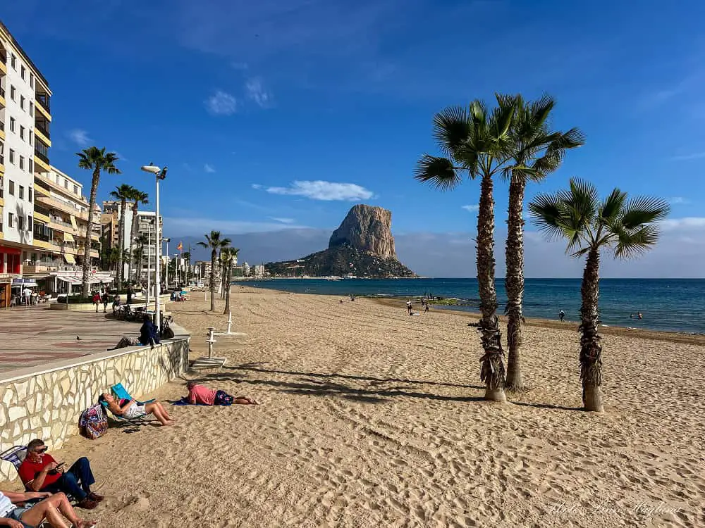 Beaches in Calpe with fine sand and palm trees. The Peñon de Ifach rock is jutting out in the sea at the far end.