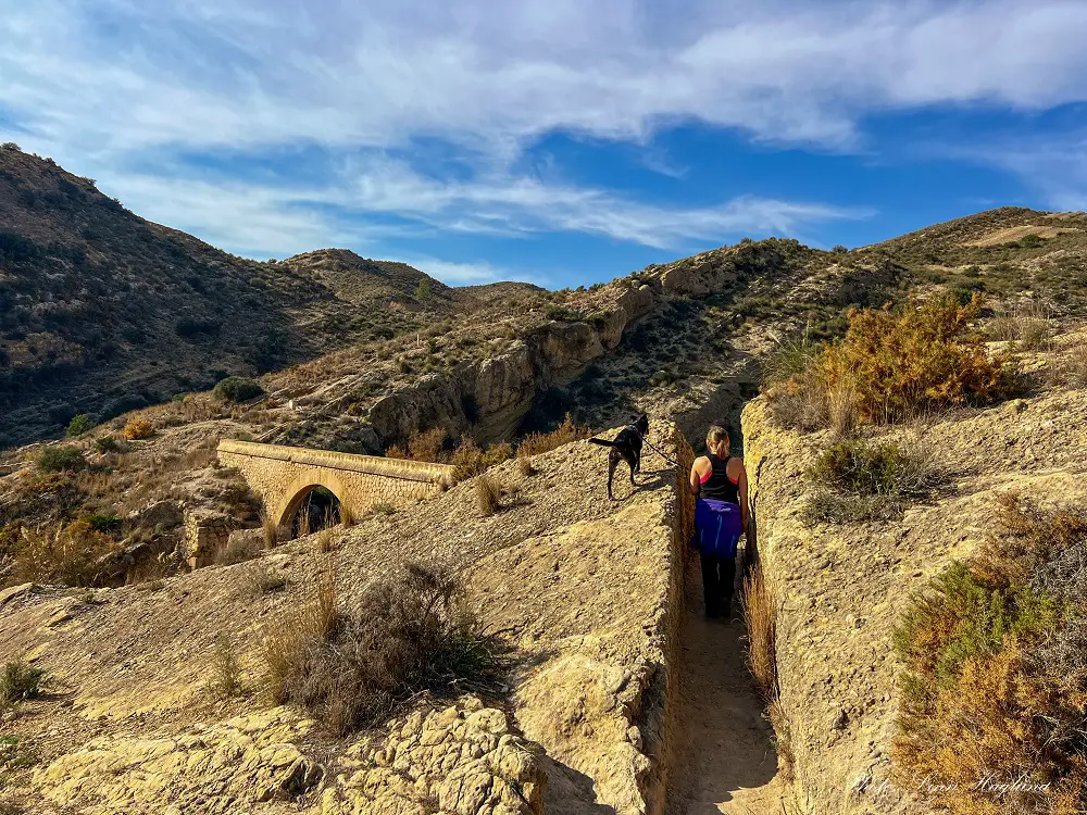 Me and Atlas walking through a narrow path cut in the rock next to an aquaduct while hiking Pantano de Elche Spain.