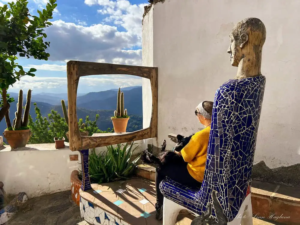 Me and my dog sitting in a mosaic chair of a human looking at beautiful views of nature through an art piece resembling a tv in one of Andalucia's hidden gems, Genalguacil.