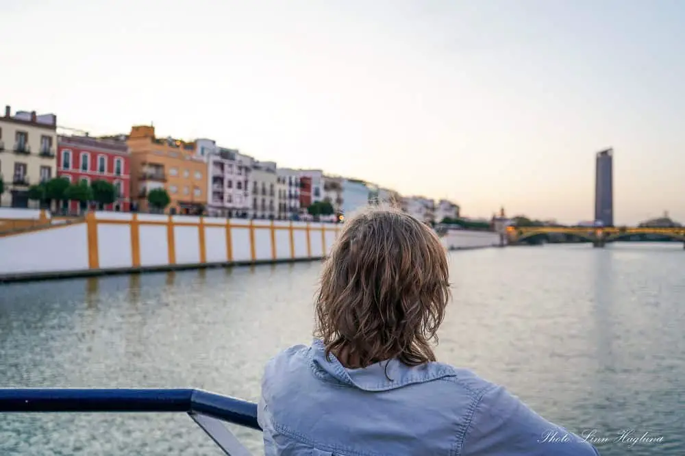 Me, in a light shirt, standing in the front of a boat, looking at the colorful Calle Betis in Triana, Seville at sunset. Going on a boat trip in Seville is one of the best things to do in the city.
