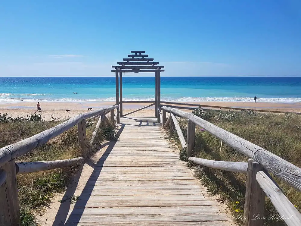 A wooden boardwalk leading to El Palmar Beach in Cadiz province in southern Spain, with a small gazebo where it meets the white-sanded beach. The day is sunny and the sea is bright blue with a few people swimming.