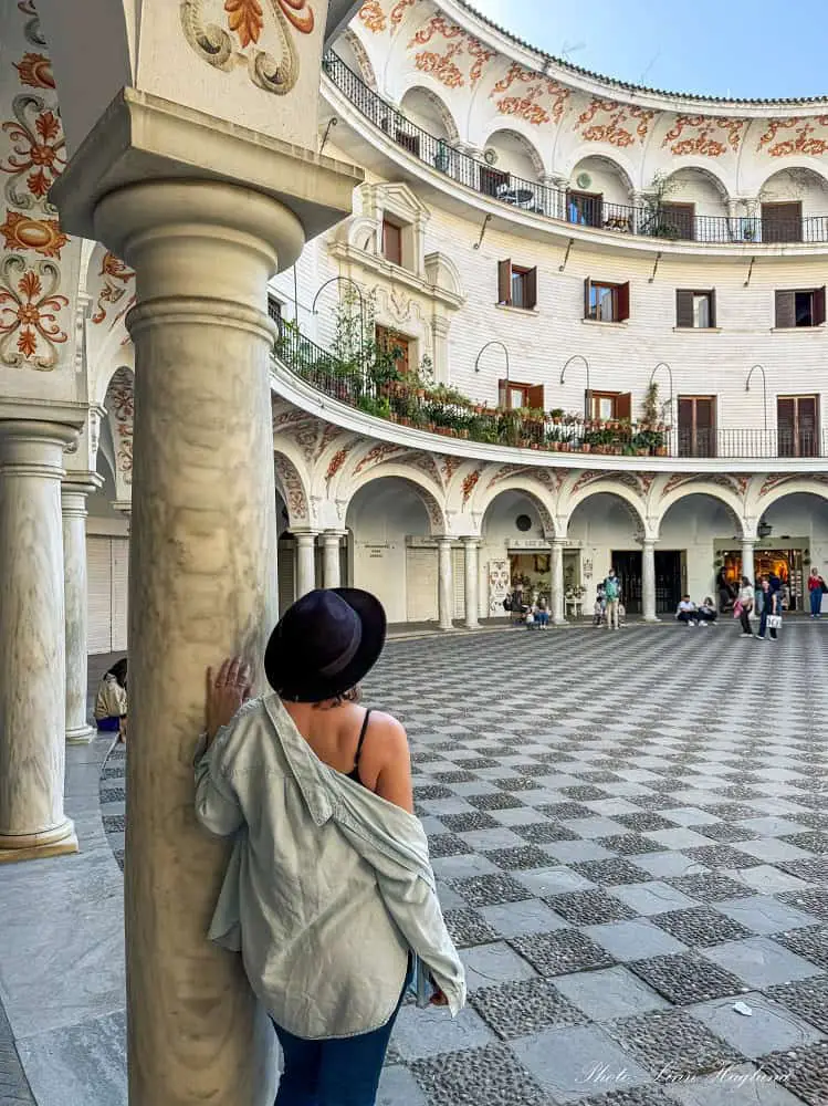 Me standing by a large pole in Plaza de Cabildo in Seville Spain. The ground is chess-shaped and there are flower-paintings on the white, oval-shaped walls.