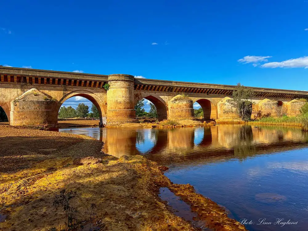 The Roman Bridge in Niebla, southern Spain, reflecting in the Rio Tinto river, which colors the riverbed and rocks orange. This is one of the most underrated day trips from Seville to take.