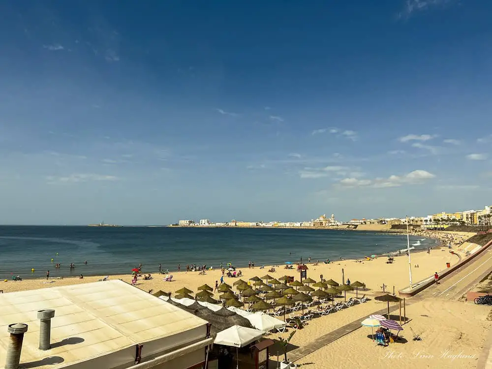 Views of Playa Santa Maria del Mar, which is one of the best beaches in Seville's surroundings. The beach is seen from the boardwalk above the beach, and parasols and a beach bar is filled with people, while the rest of the white-sanded beach that leads to Cadiz city only has a few people walking on it.