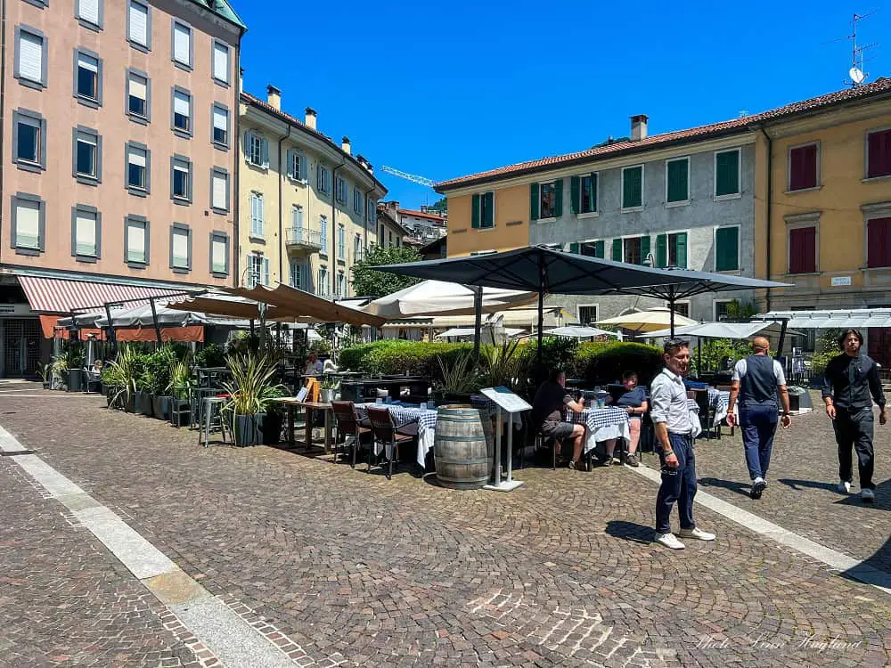 People walking around a square with restaurants, pot plants, and pastel colored houses. This is a lovely place to walk around when visiting Como in one day.