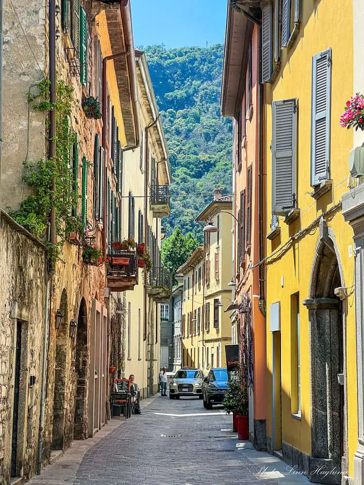 A narrow street in Como Old Town with pastel colored houses and mountain views at the end of the street. Lush plants cascade out of rustic balconies and windows soaking up the sun.