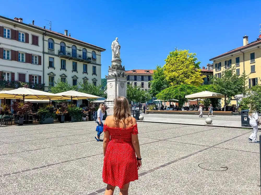 Me in a red dress standing in the middle of Piazza Alessandro Volta in Como looking at the statue of the scientist by the same name. People watching in this square is one of the top things to do in Como Italy.