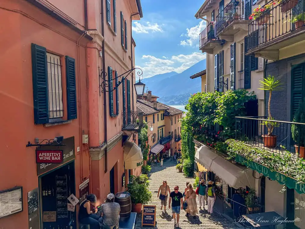 People walking up the steep, stepped street, Salita Serbelloni in Bellagio, where there are boutiques, bars, and restaurants. The pastel-colored houses lead to lake and mountain views of Lake Como, Italy.