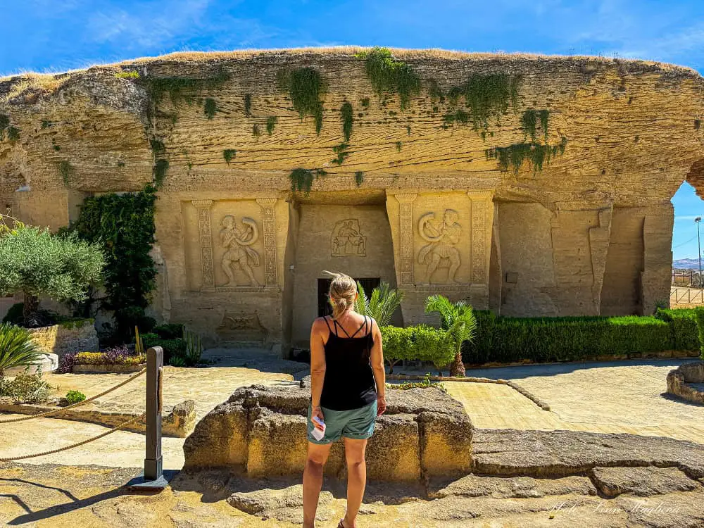 Me, in shorts and singlet, looking at the entrance of a large rock with giant relics on each side of a central door. This is the Coto de las Canteras in Osuna Spain, which is also known as the Petra of Spain.